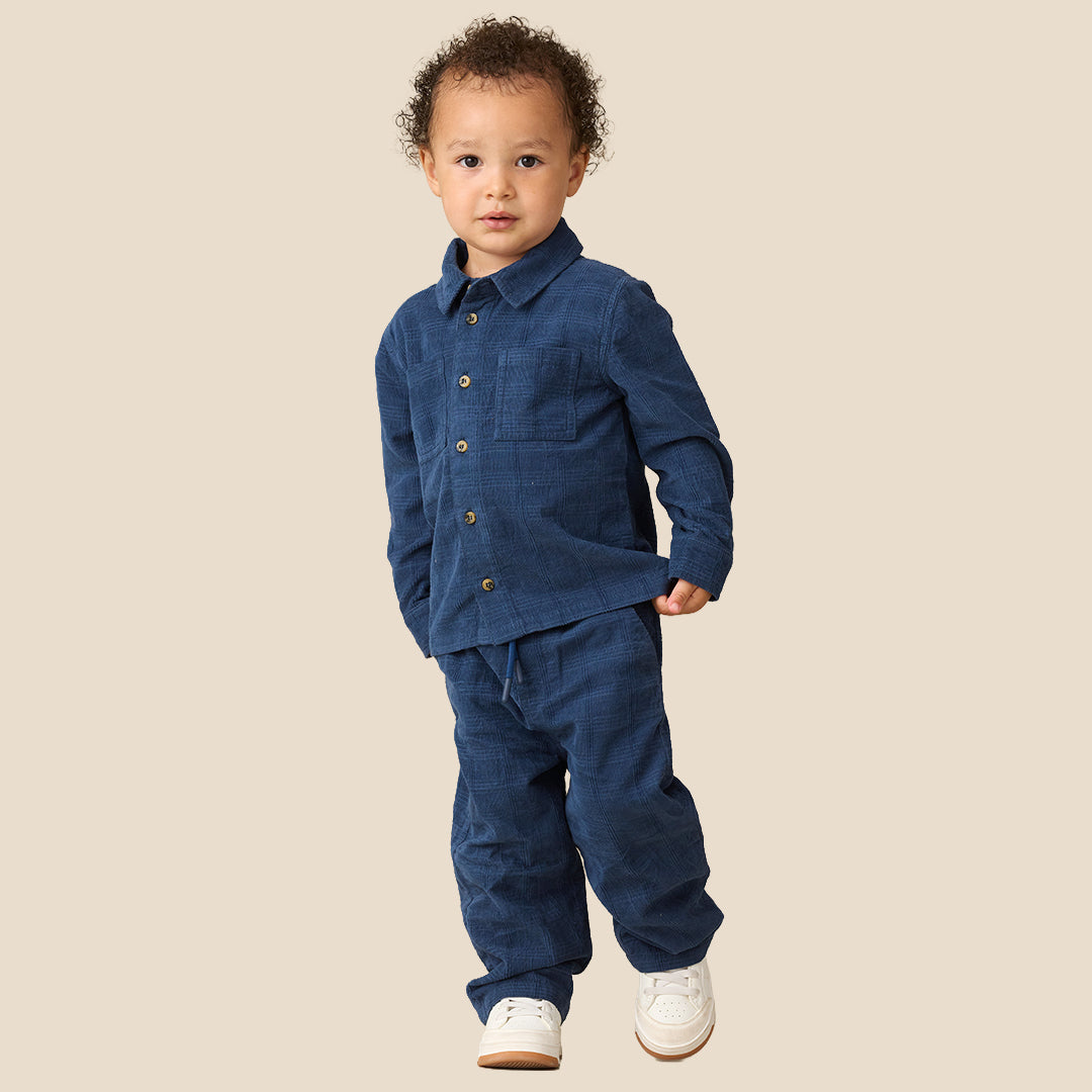 A toddler standing against a plain beige studio background, wearing a matching dark blue denim-like two-piece outfit with a collared long-sleeve top and pants, plus white sneakers.