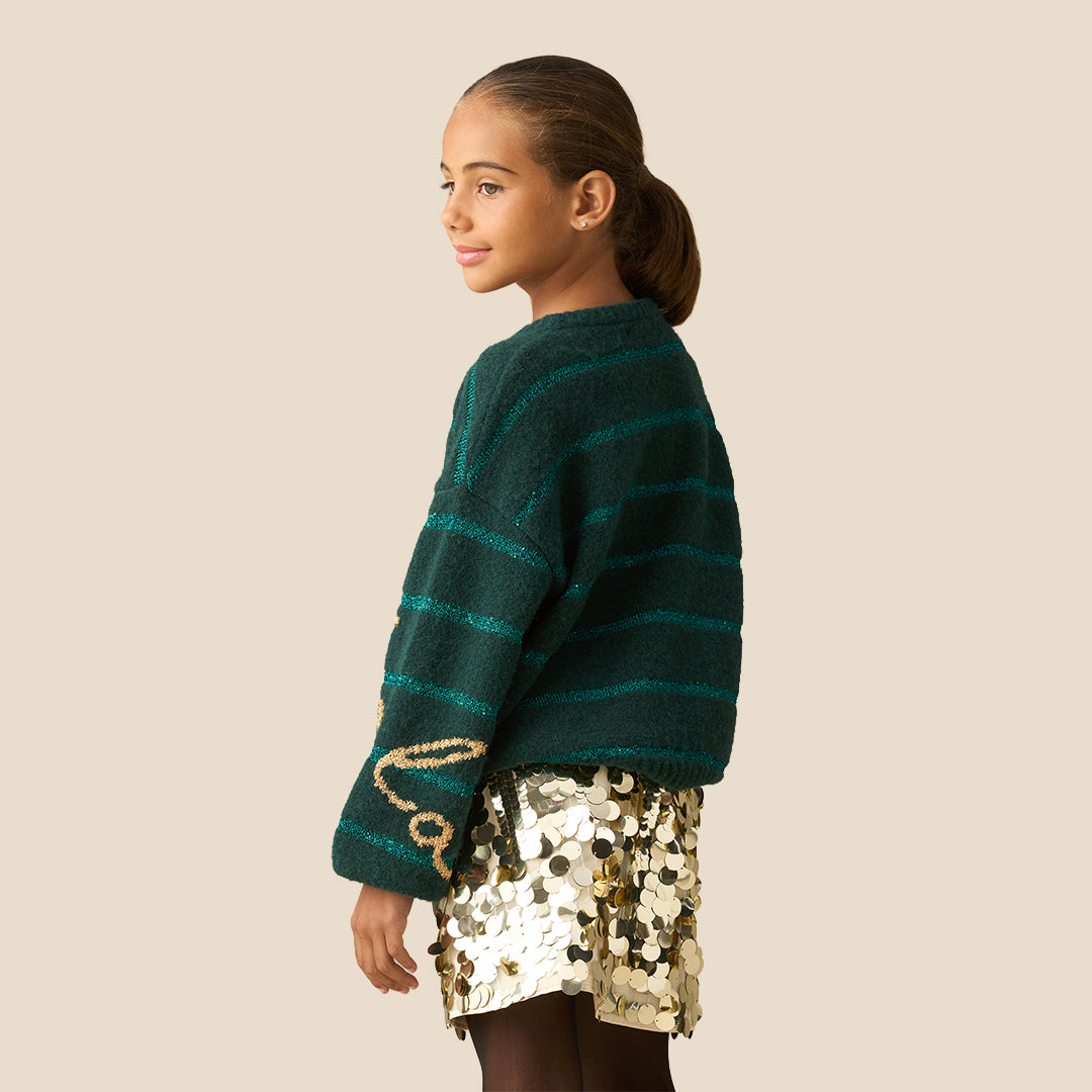 A young girl smiling while wearing a dark green knit sweater with teal horizontal stripes and gold glittery 'fo'lo' lettering on the front, paired with a gold sequined skirt, against a solid beige background.