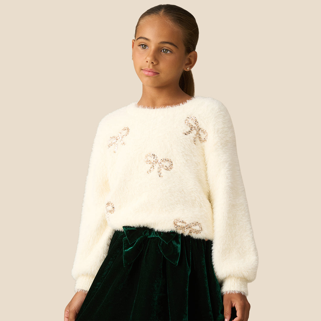 A young girl with her hair in a ponytail wearing a fuzzy off-white long-sleeve sweater with sequined bow details and a dark green velvet skirt, set against a solid beige background.