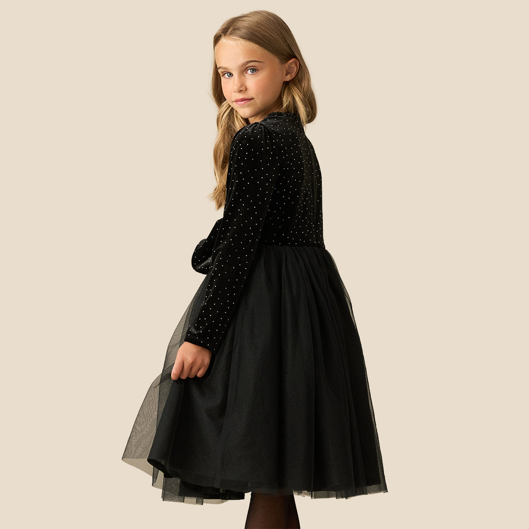 A young girl with blonde hair wearing a black long-sleeve dress with a polka-dot velvet bodice, a sheer black tulle skirt, and a matching bow belt, posing against a solid beige background.