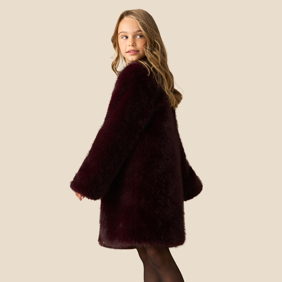 A young girl with blonde hair wearing a plush, dark burgundy faux fur coat over a dark patterned dress, standing against a solid beige background.