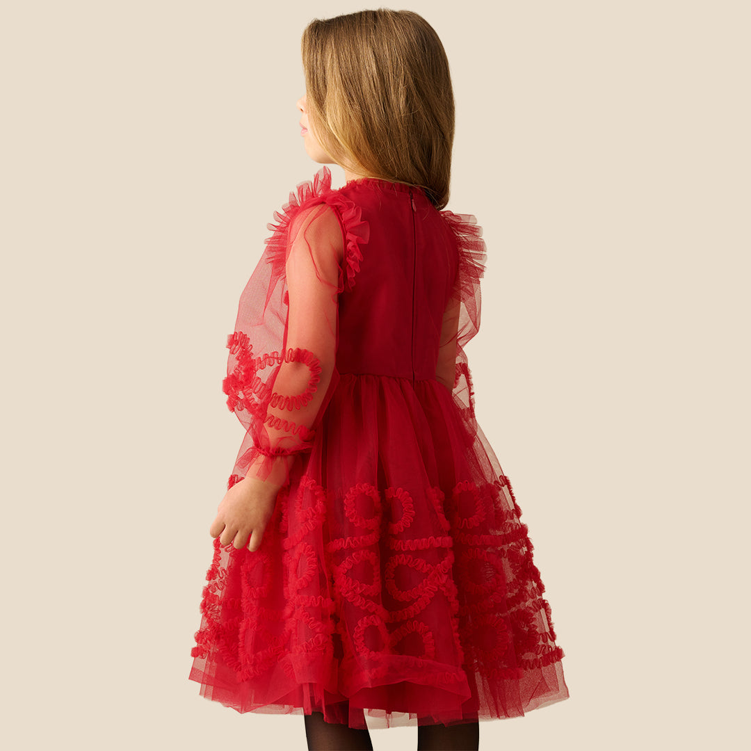 A young girl with blonde hair wearing a red tulle dress with red lace appliques and sheer flutter sleeves, standing against a solid beige background.