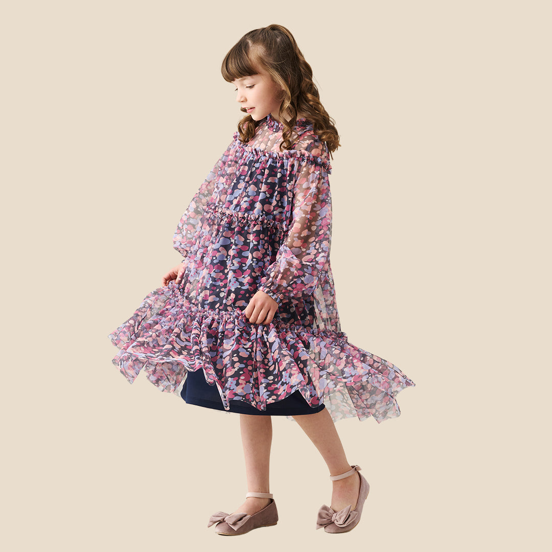 A young girl with curly hair stands on a checkered porch wearing a long-sleeved tiered dress with a pink, purple, and blue floral pattern. She is posing behind a black wrought-iron fence.