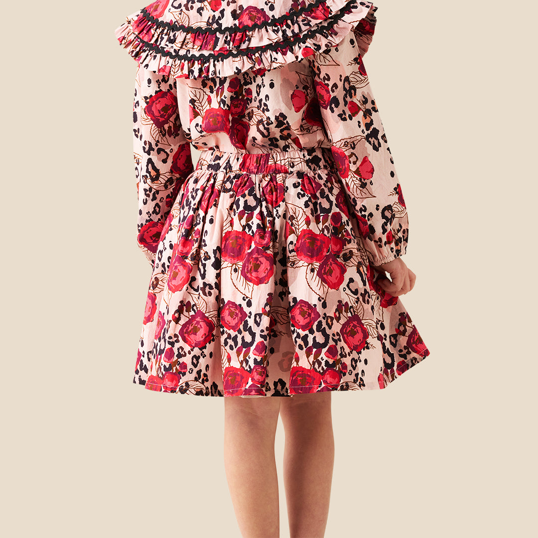 A young girl wearing a long-sleeve dress with a red rose and black leopard print pattern on a light pink background, photographed against a solid beige backdrop.