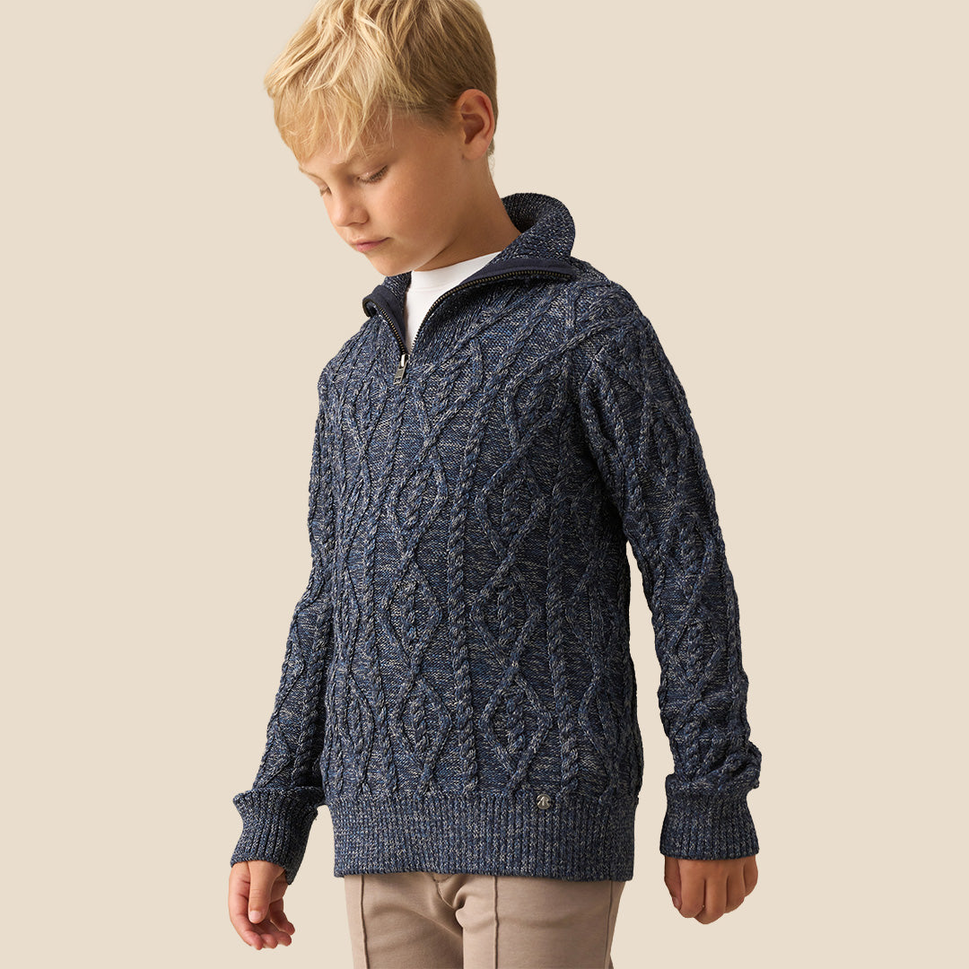 A young boy with blonde hair wearing a navy blue cable-knit mock-neck sweater and beige trousers, standing against a solid beige background.