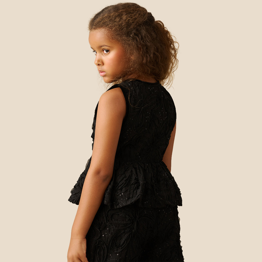 A young girl with curly brown hair wearing a sleeveless black textured dress with a ruffled peplum and bow detail, posing against a solid beige background.