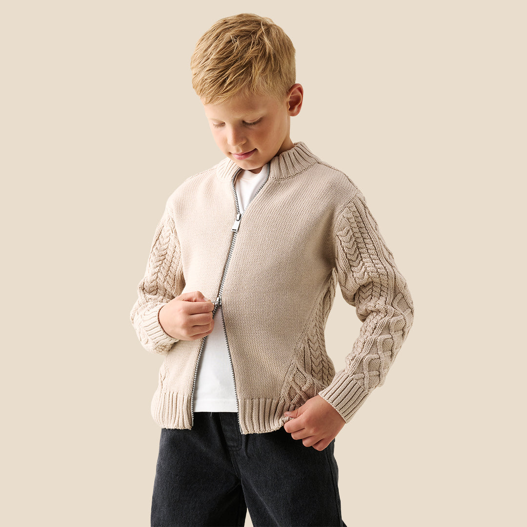 A young boy wearing a beige cable-knit zip-up cardigan over a white t-shirt and dark jeans, standing against a solid light beige background.