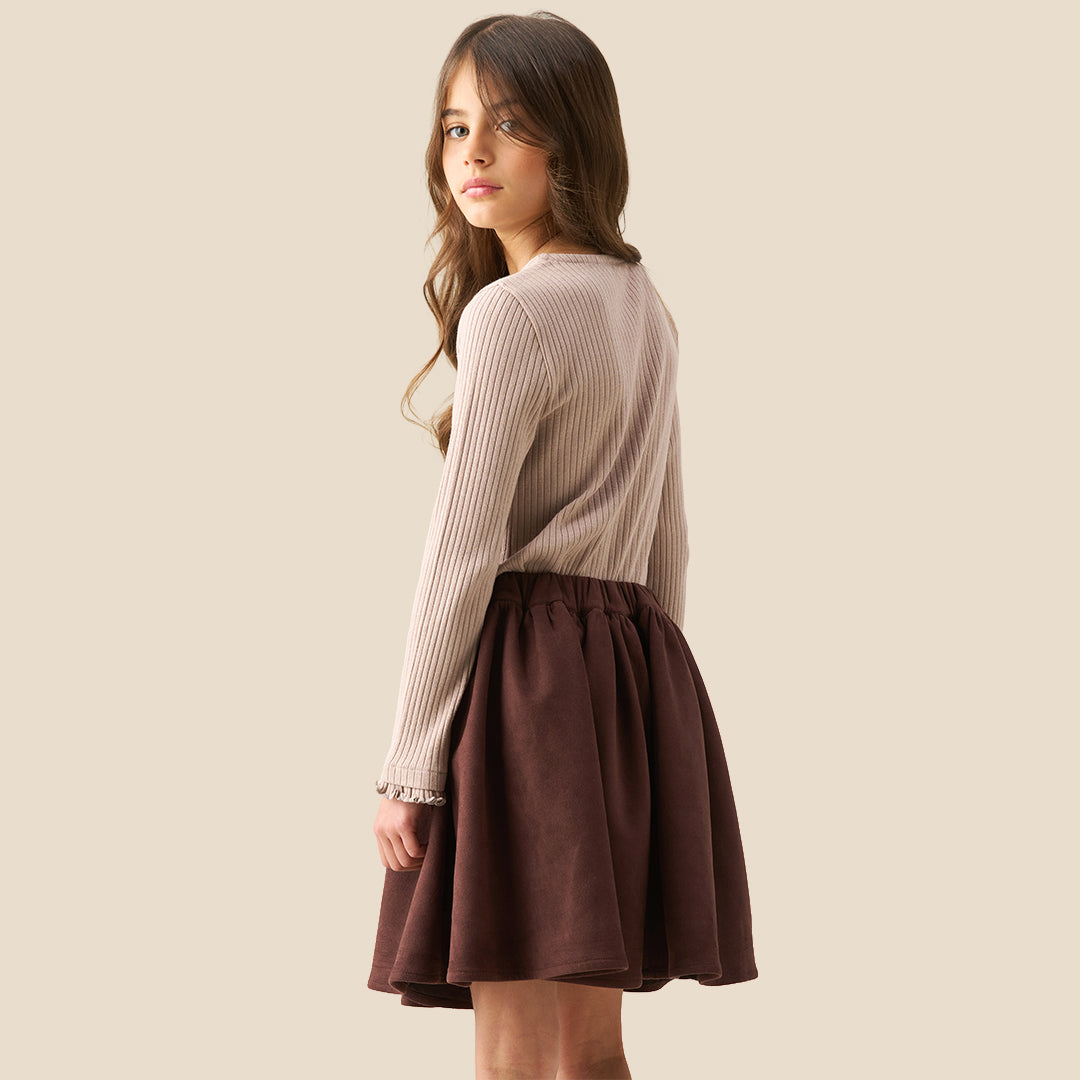 A model wearing a beige ribbed cardigan and a dark brown pleated skirt with a large bow at the waist, shown from the chest down against a solid beige background.