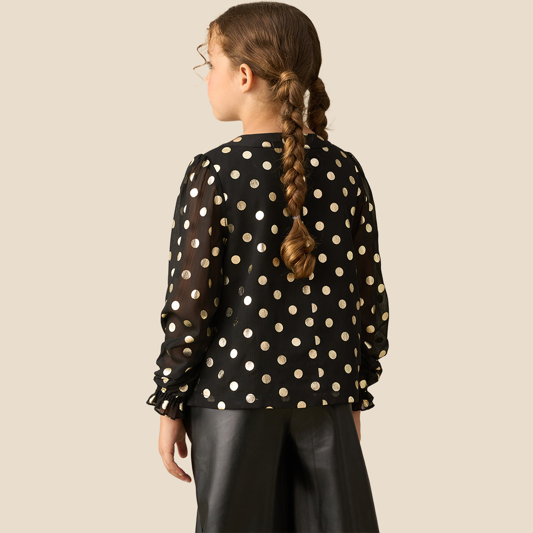 A young girl with braided hair wearing a black long-sleeve blouse with gold polka dots and black faux-leather pants, standing against a solid beige background.