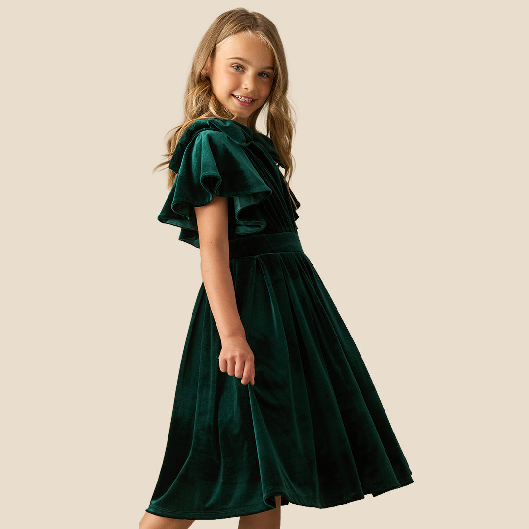 A young girl wearing a dark green velvet dress with ruffled short sleeves, a matching waist belt, and a pleated skirt, standing against a solid beige background.