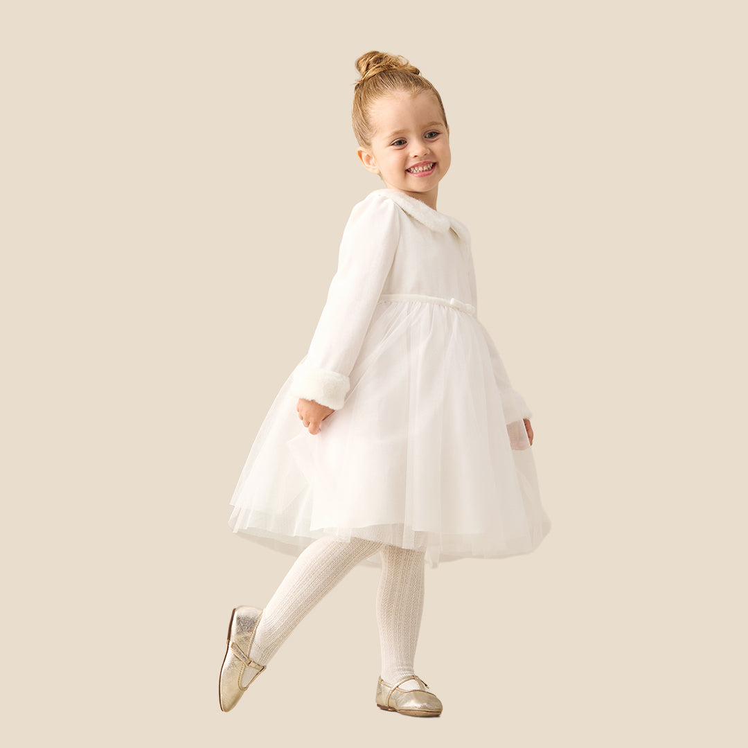 A young girl with blonde hair in a bun wearing a white long-sleeve tulle dress with faux fur trim on the collar and cuffs, standing against a solid beige background.