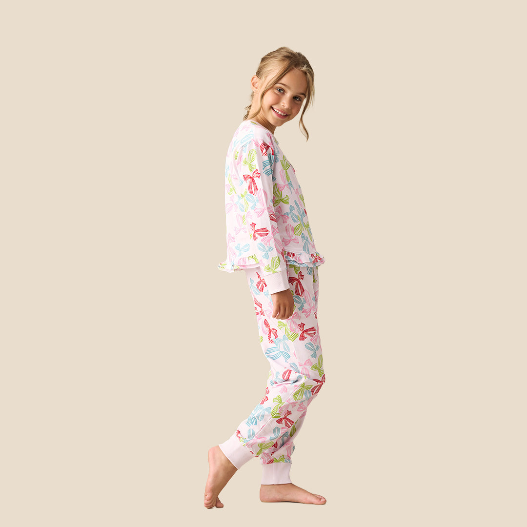 A young girl modeling a two-piece pajama set featuring a long-sleeve top and matching pants with a colorful bow pattern on a light pink background, set against a solid beige backdrop.