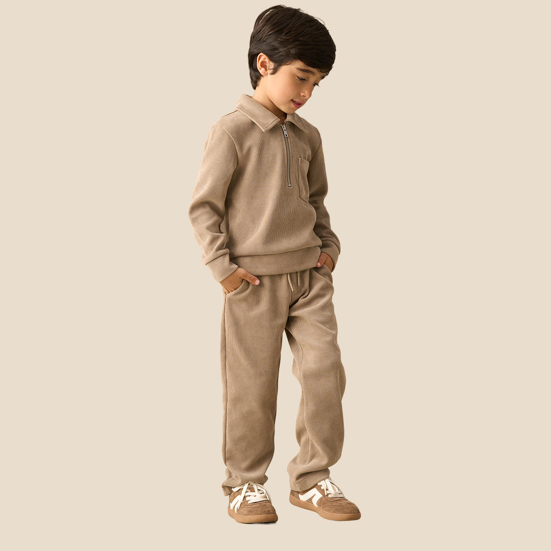 A child wearing a matching beige fleece tracksuit with a half-zip sweatshirt and relaxed-fit pants, paired with brown sneakers, against a solid light beige background.