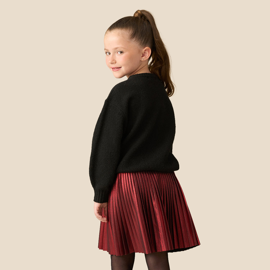 A young girl with her hair in a ponytail is wearing a black knit sweater featuring a large bow design outlined with colorful Christmas string lights, paired with a red pleated skirt, set against a solid beige background.