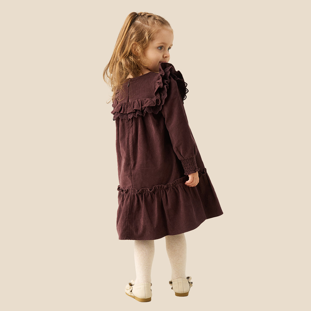 A young girl with light brown hair in pigtails stands against a solid beige background wearing a long-sleeved dark plum-brown dress with ruffles, white tights, and cream-colored shoes.