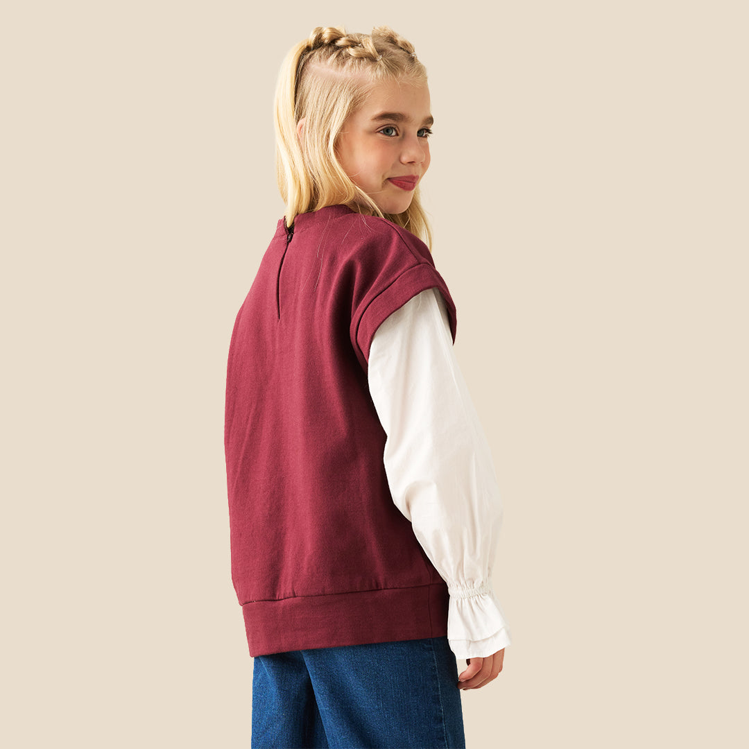 A young girl with blonde pigtails wearing a maroon cropped t-shirt with white sleeves and 'C'EST LA VIE paris' printed in white, layered over a white long-sleeve blouse with ruffled cuffs, and blue jeans, against a solid beige background.