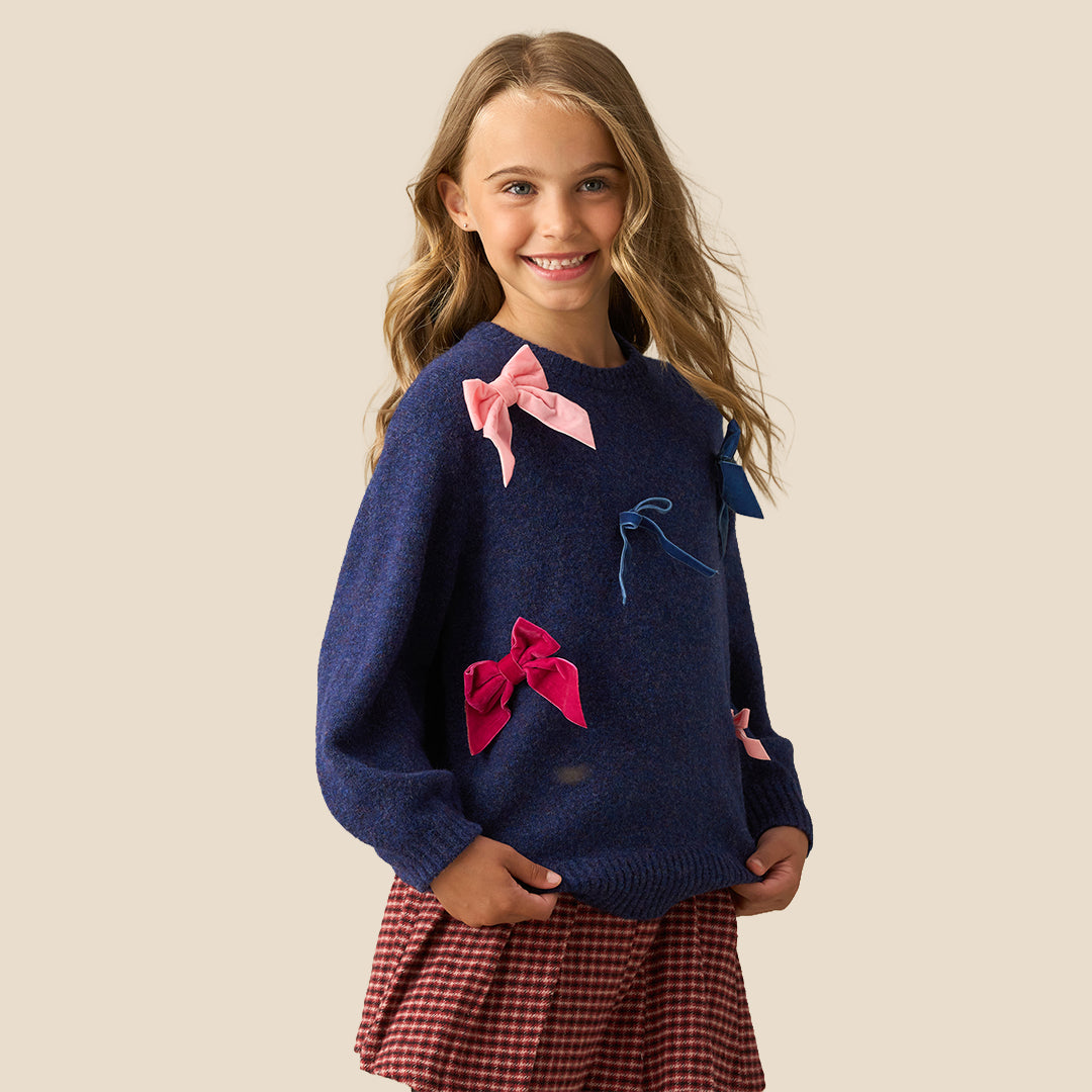 A young girl with blonde hair is smiling and posing in a navy blue knit sweater adorned with pink and blue fabric bows, paired with a red and black gingham pleated skirt, set against a solid beige background.