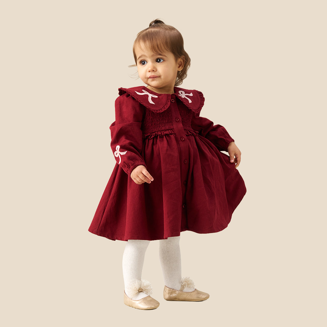 A toddler girl wearing a burgundy corduroy dress with a large white ruffled collar decorated with bows, white tights, and beige ballet flats, standing against a solid beige background.