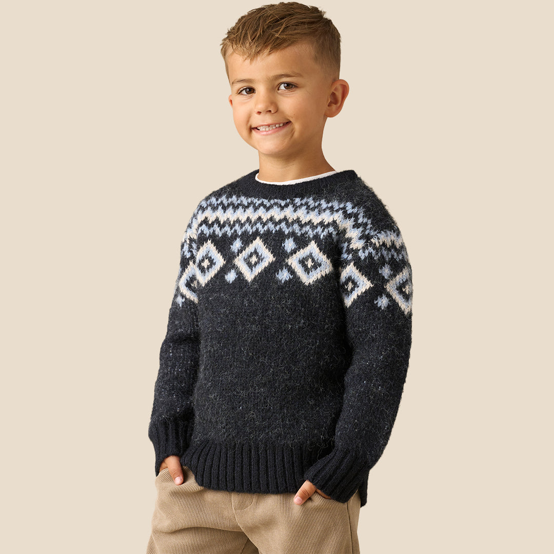 A young boy wearing a dark navy knit sweater with a white and light blue geometric Fair Isle pattern across the chest and shoulders, paired with beige pants, set against a solid light beige background.