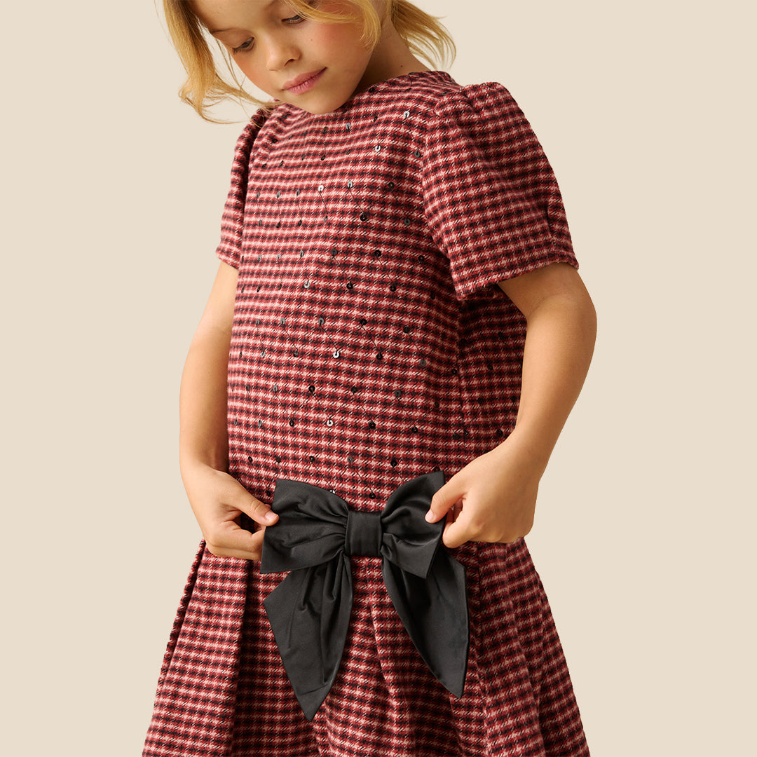A young girl smiling and posing in a red and white gingham dress with a large black bow at the waist, set against a solid beige background.
