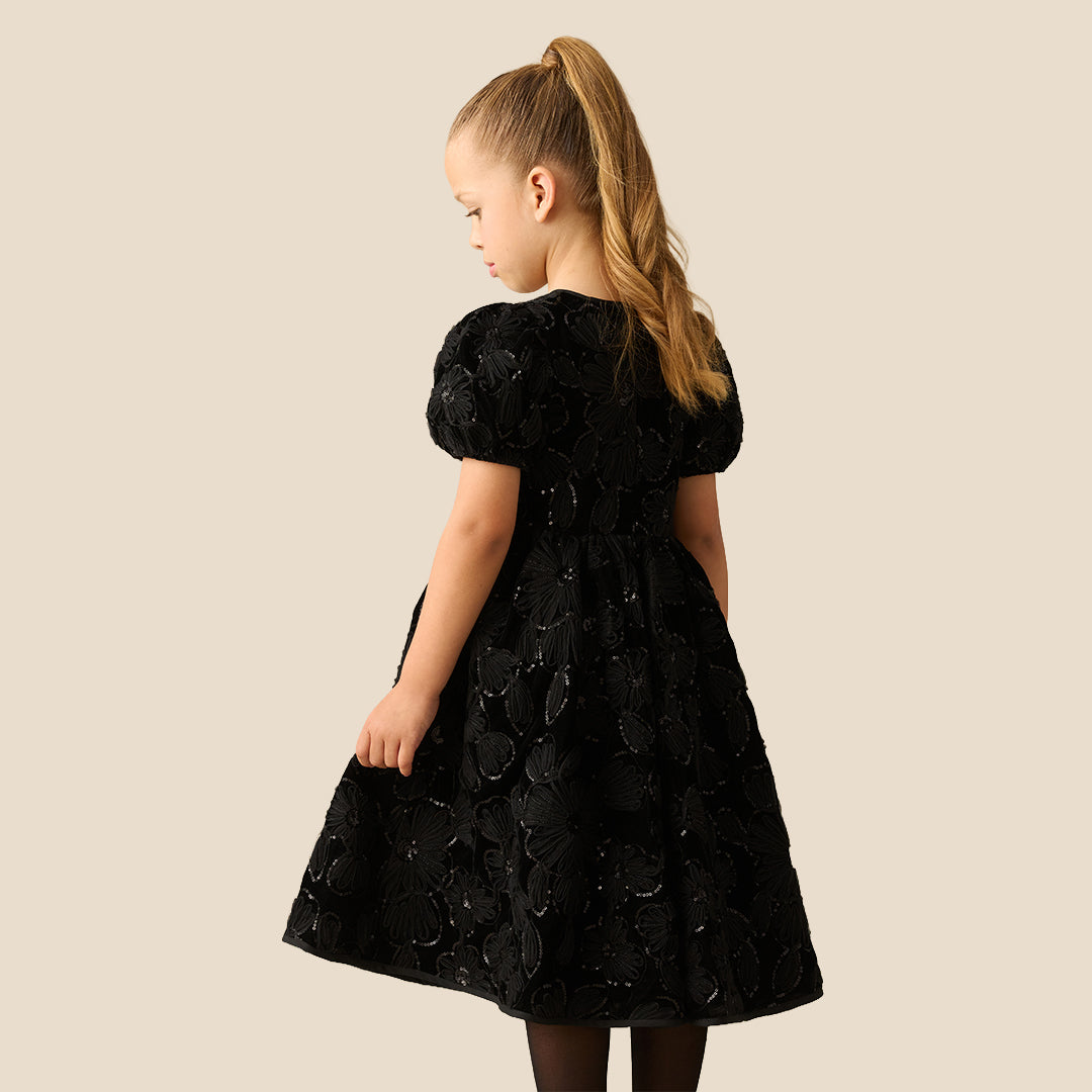 A young girl with her hair in a ponytail models a black short-sleeve dress with textured floral applique, standing against a solid beige background.