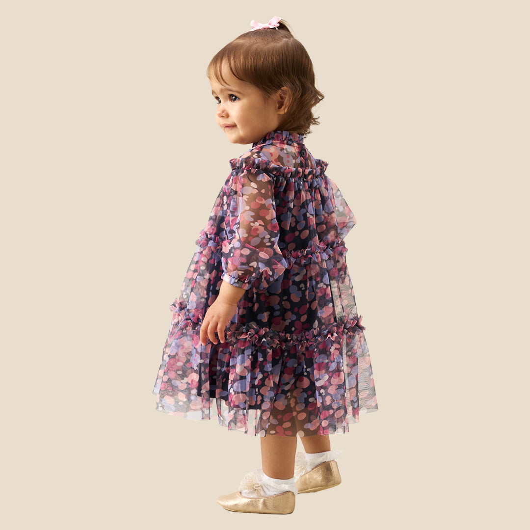 A toddler girl wearing a tiered long-sleeve dress with a dark navy base and a colorful floral pattern in pink, purple, and blue, standing against a solid beige background.