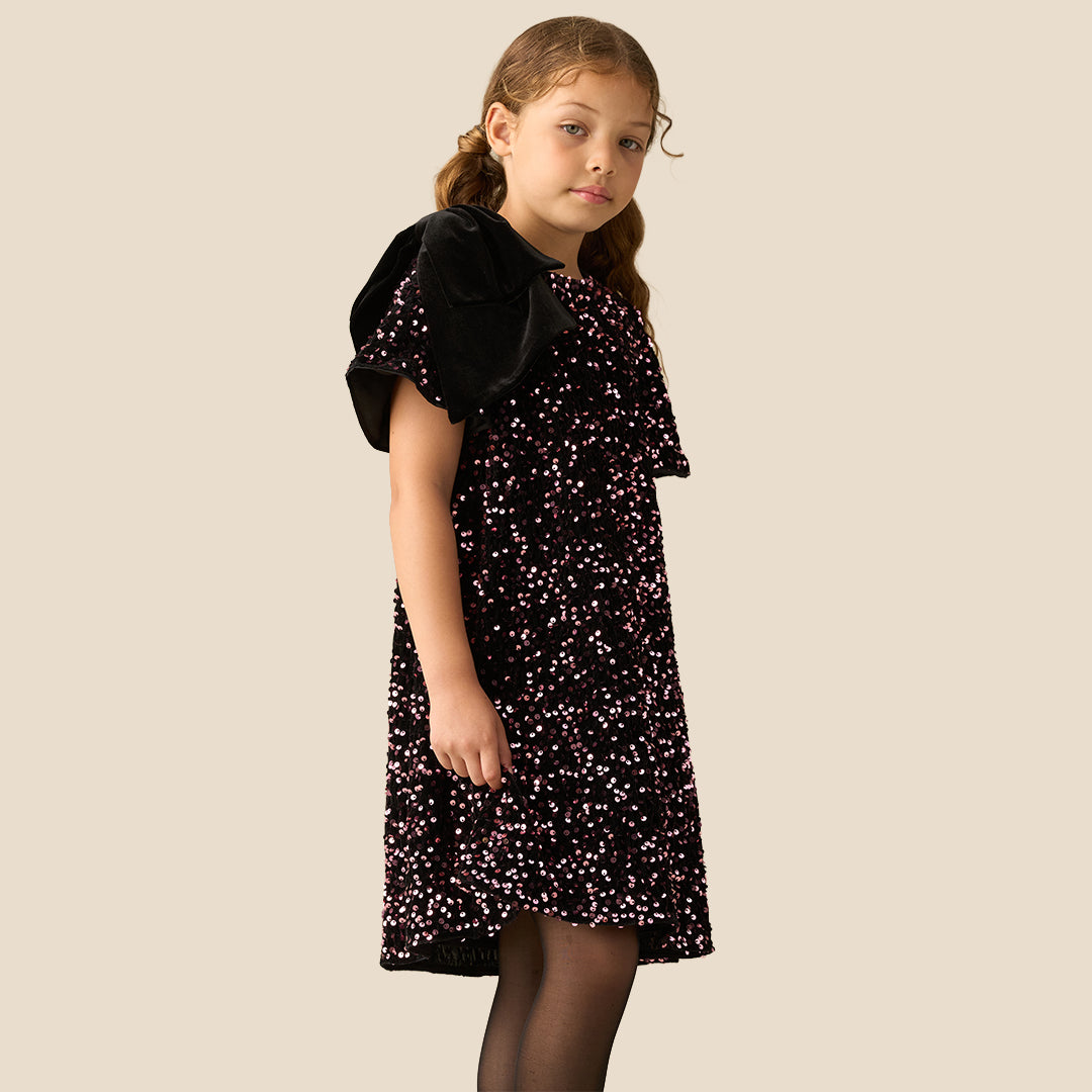 A young girl with light brown hair in pigtails stands against a solid beige background wearing a short-sleeved black dress covered in small pink and white floral patterns, with a black bow on the shoulder.
