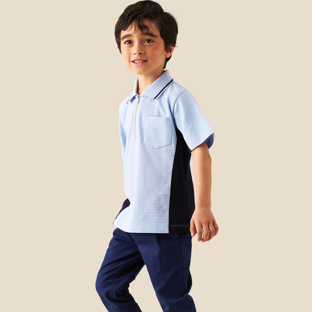 A young boy modeling a light blue polo shirt with dark navy side panels and a half-zip collar, paired with navy pants, against a solid beige background.