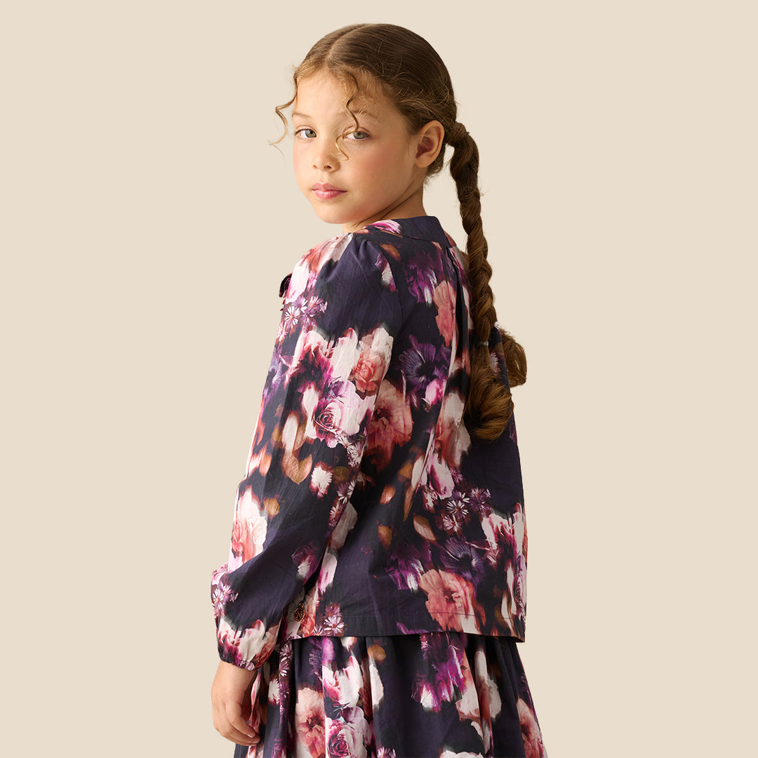 A young girl with braided hair wearing a long-sleeve floral blouse with a large bow at the collar, photographed against a plain light beige studio background.