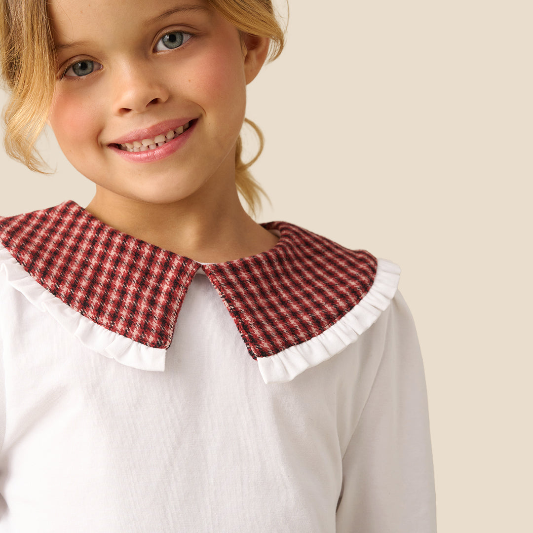 A young girl smiling and looking to the side, wearing a white long-sleeve blouse with a red and black gingham collar, paired with a matching red and black gingham pleated skirt, against a solid beige background.
