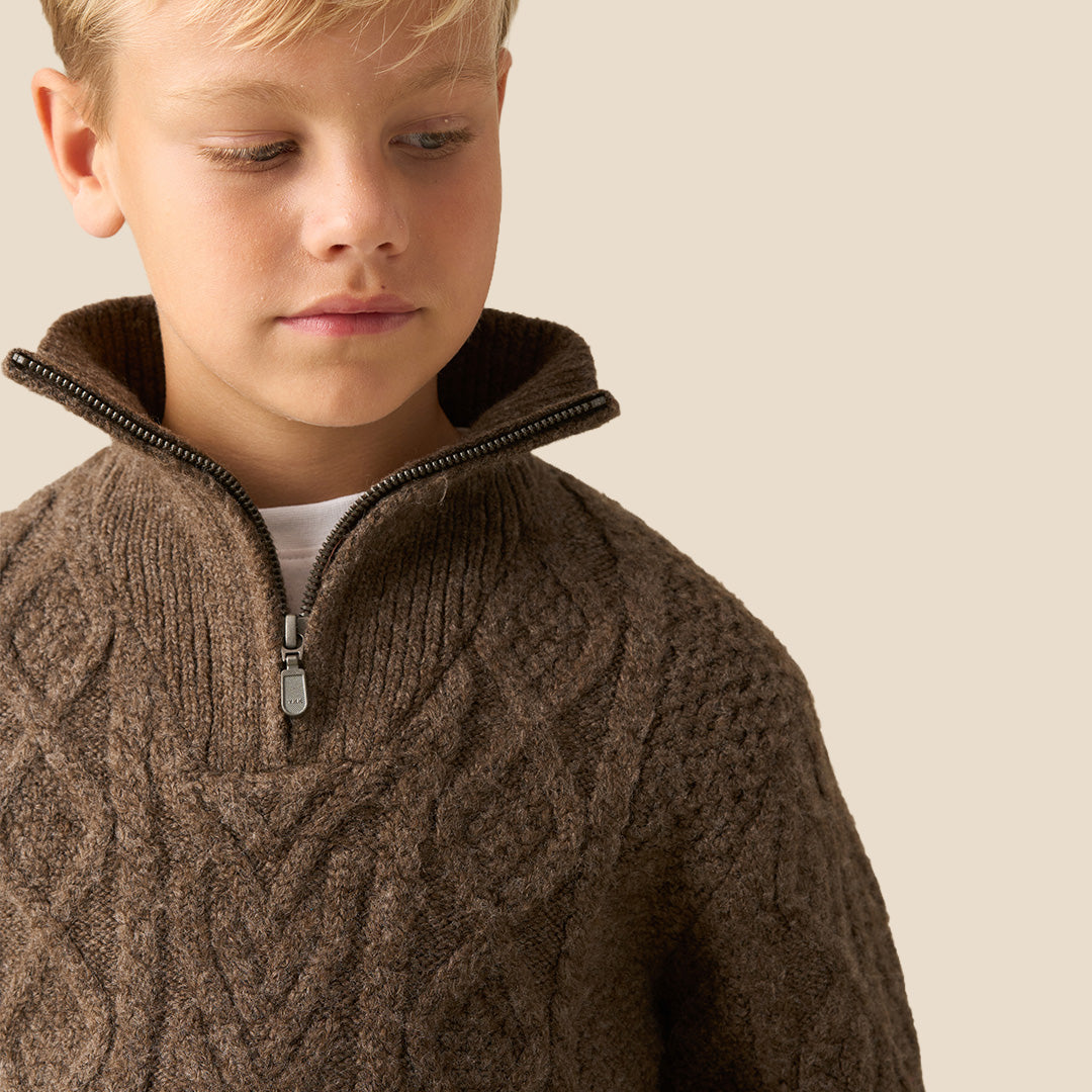 A child wearing a brown cable-knit mock-neck sweater and light pink trousers, standing indoors near a white wall and window. Another child in a brown-and-white plaid shirt is partially visible in the foreground.