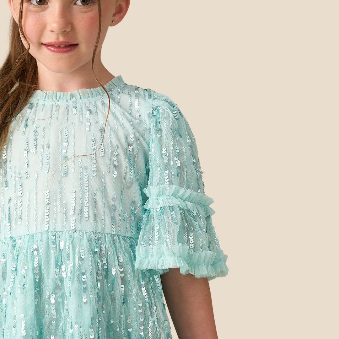 A young girl with a long brown ponytail wearing a light mint green sequined tiered dress, standing against a solid beige background.