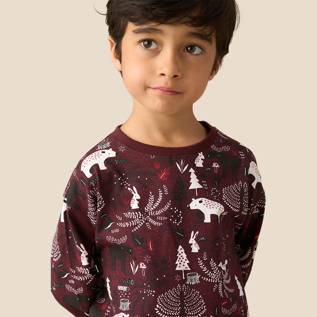 A young boy wearing a matching long-sleeve pajama set with a woodland animal print, standing against a solid light beige background.