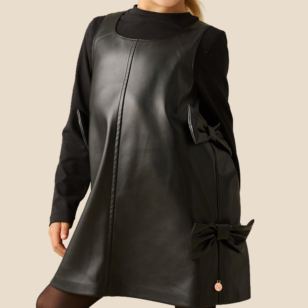 A young girl with blonde hair tied back is wearing a black faux-leather dress with long sleeves, a mock neck, and decorative bows on the sides, paired with black tights, against a solid beige background.