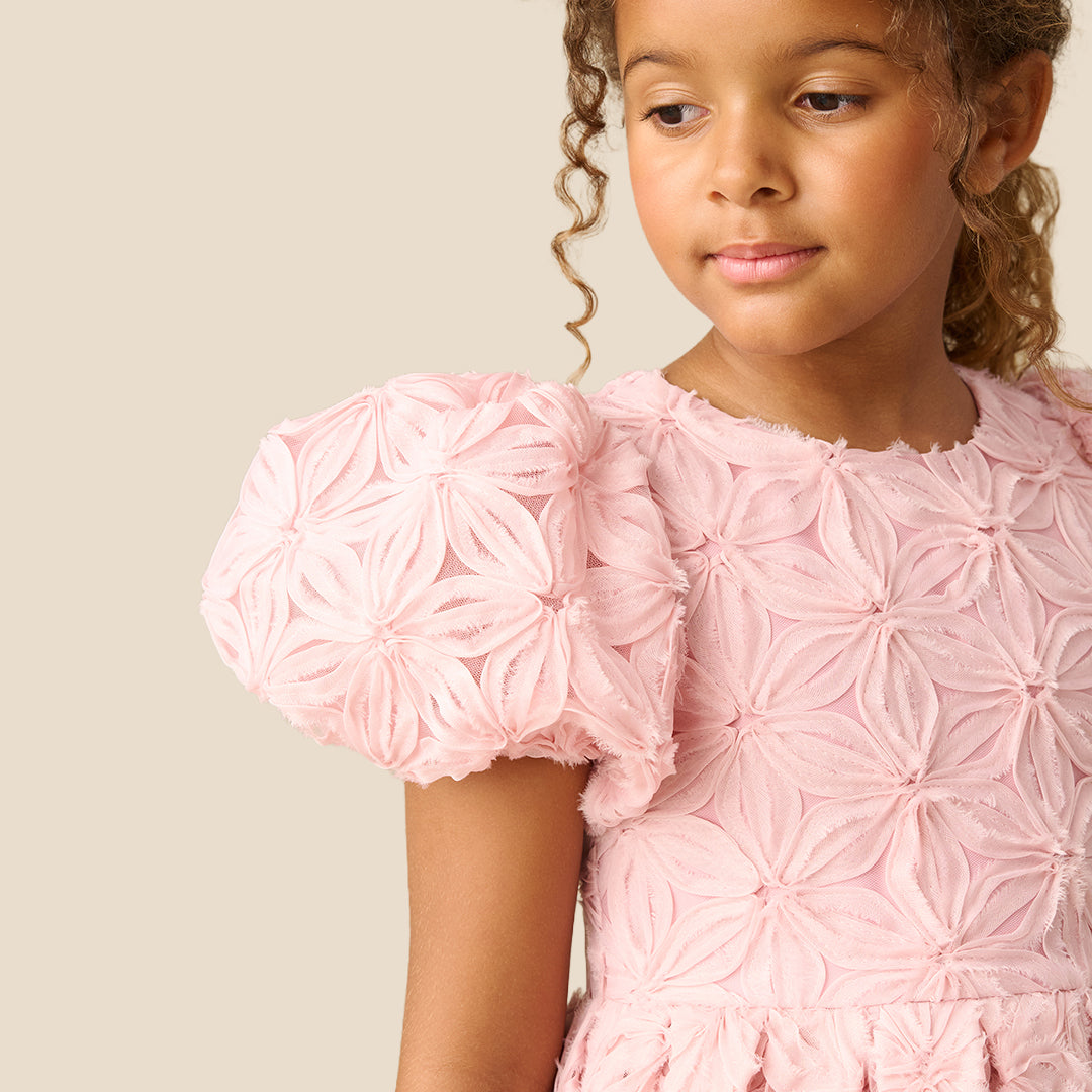 A young girl with curly brown hair wearing a light pink textured dress with puffed short sleeves and a full skirt, standing against a solid beige background.