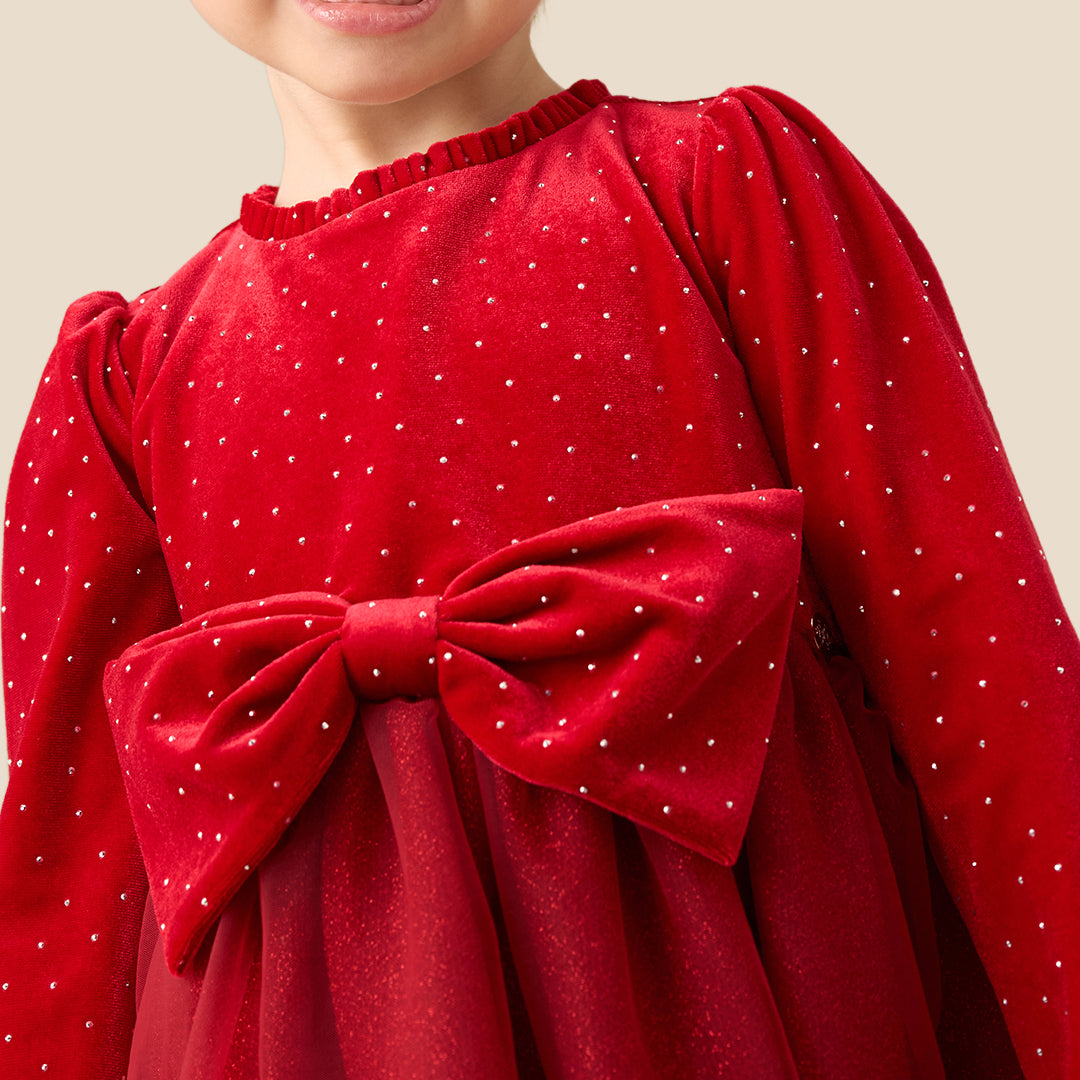 A young girl with her hair in a bun stands against a solid beige background wearing a festive red dress with a polka-dot velvet bodice, long sleeves, a large matching bow at the waist, and a tulle skirt. She is also wearing white knit tights and metallic gold shoes.