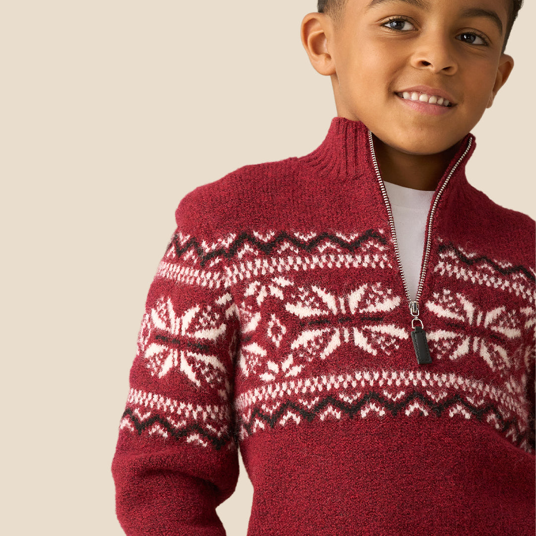 A young boy smiling and wearing a burgundy knit quarter-zip sweater with a white and dark red Fair Isle snowflake pattern across the chest and shoulders, set against a solid beige background.