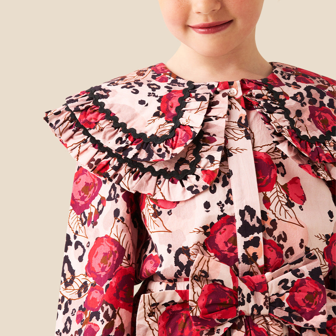 A young girl with her hair in a bun stands wearing a long-sleeve dress with a red rose and black leopard print pattern on a light pink background, set against a solid beige studio backdrop.