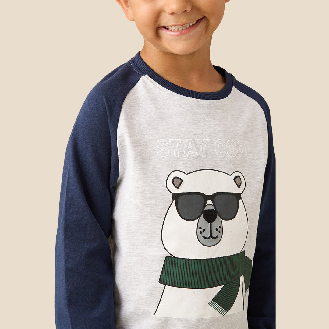 A young boy modeling a two-piece pajama set against a solid beige background. The set features a white long-sleeve top with navy blue raglan sleeves and a cartoon bear graphic wearing sunglasses and a green scarf, paired with navy blue pants decorated with patchwork-style graphic details.