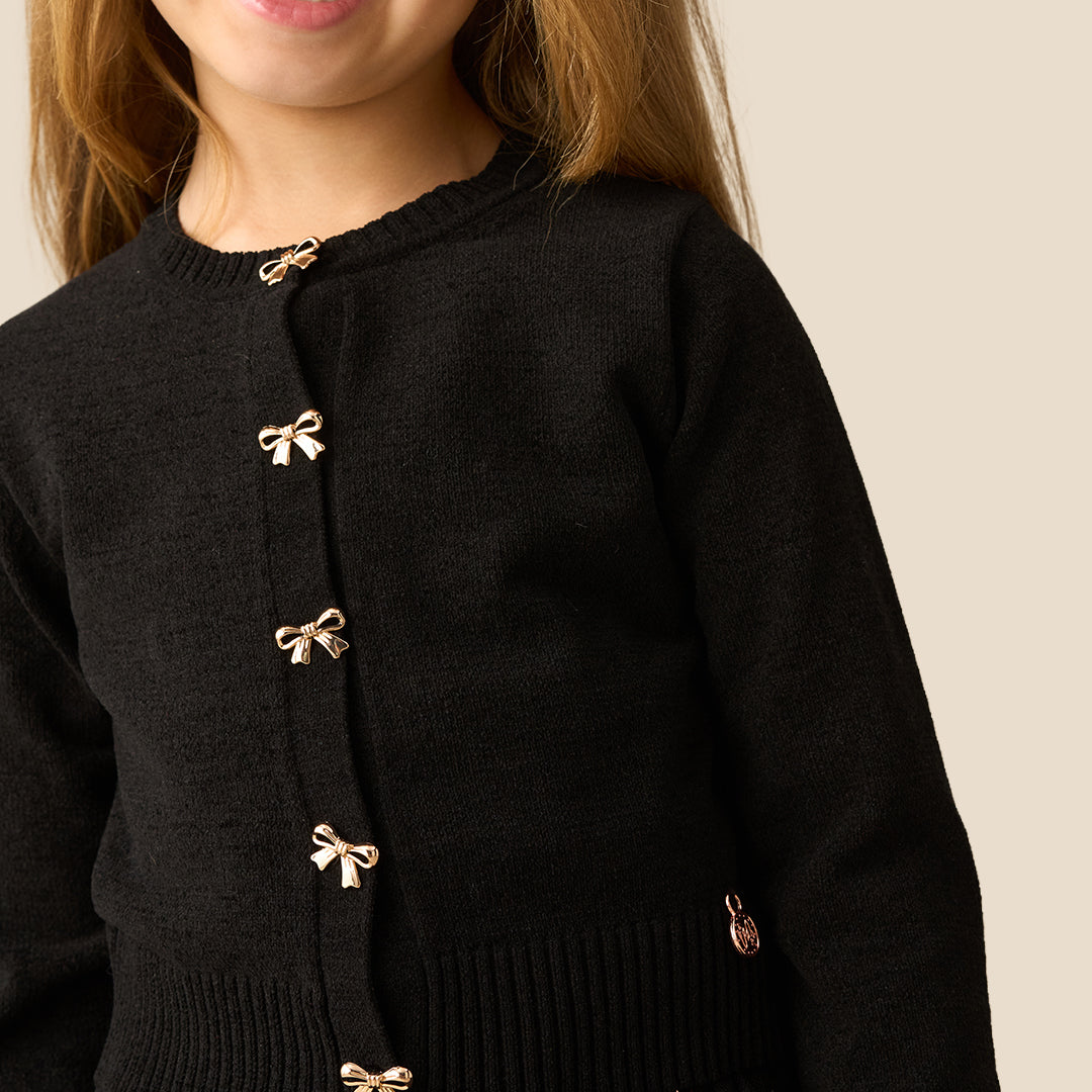A young girl with long light brown hair wearing a black knit cardigan with gold bow-shaped buttons and a black tulle skirt, standing against a solid beige background.