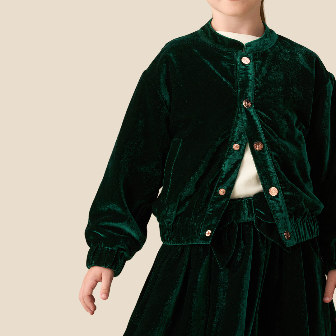 A young girl with her hair in a ponytail wearing a dark green velvet jacket and matching skirt over a white top, photographed against a solid beige studio background.