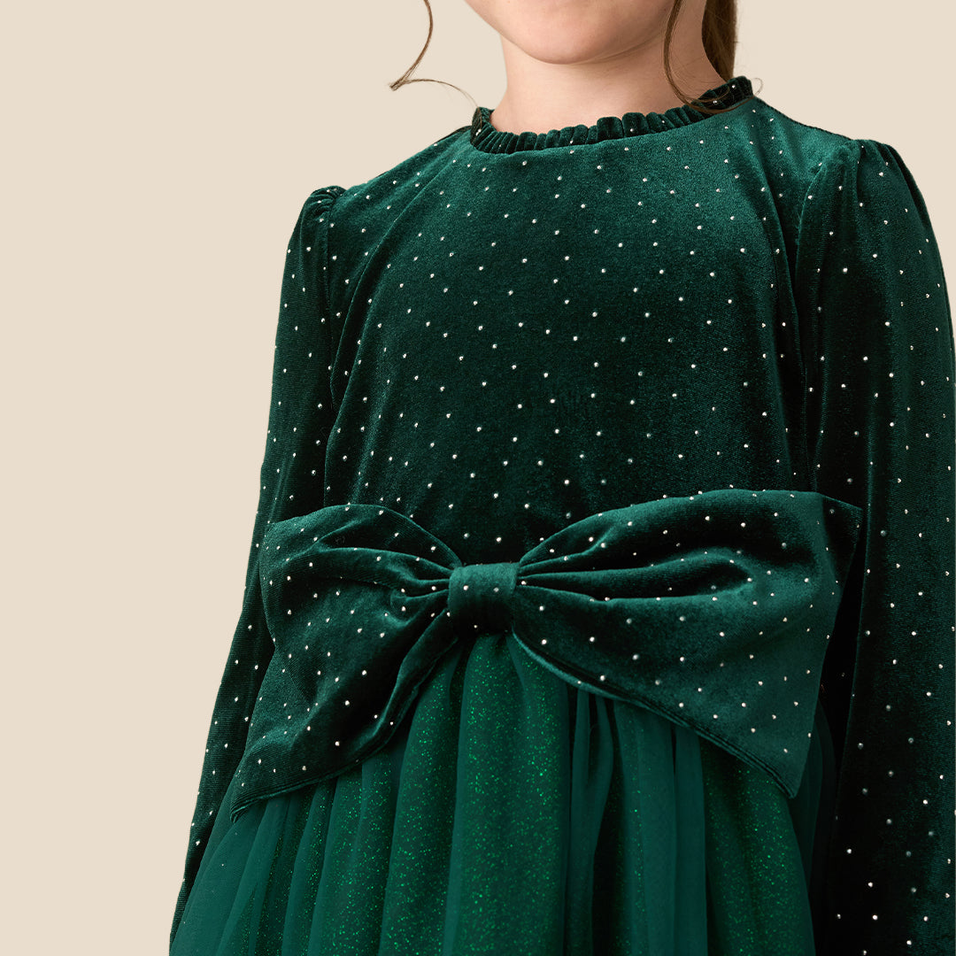 A young girl wearing a dark green velvet long-sleeve dress with a polka-dot pattern, a matching tulle skirt, and a large bow at the waist, standing against a solid beige background.