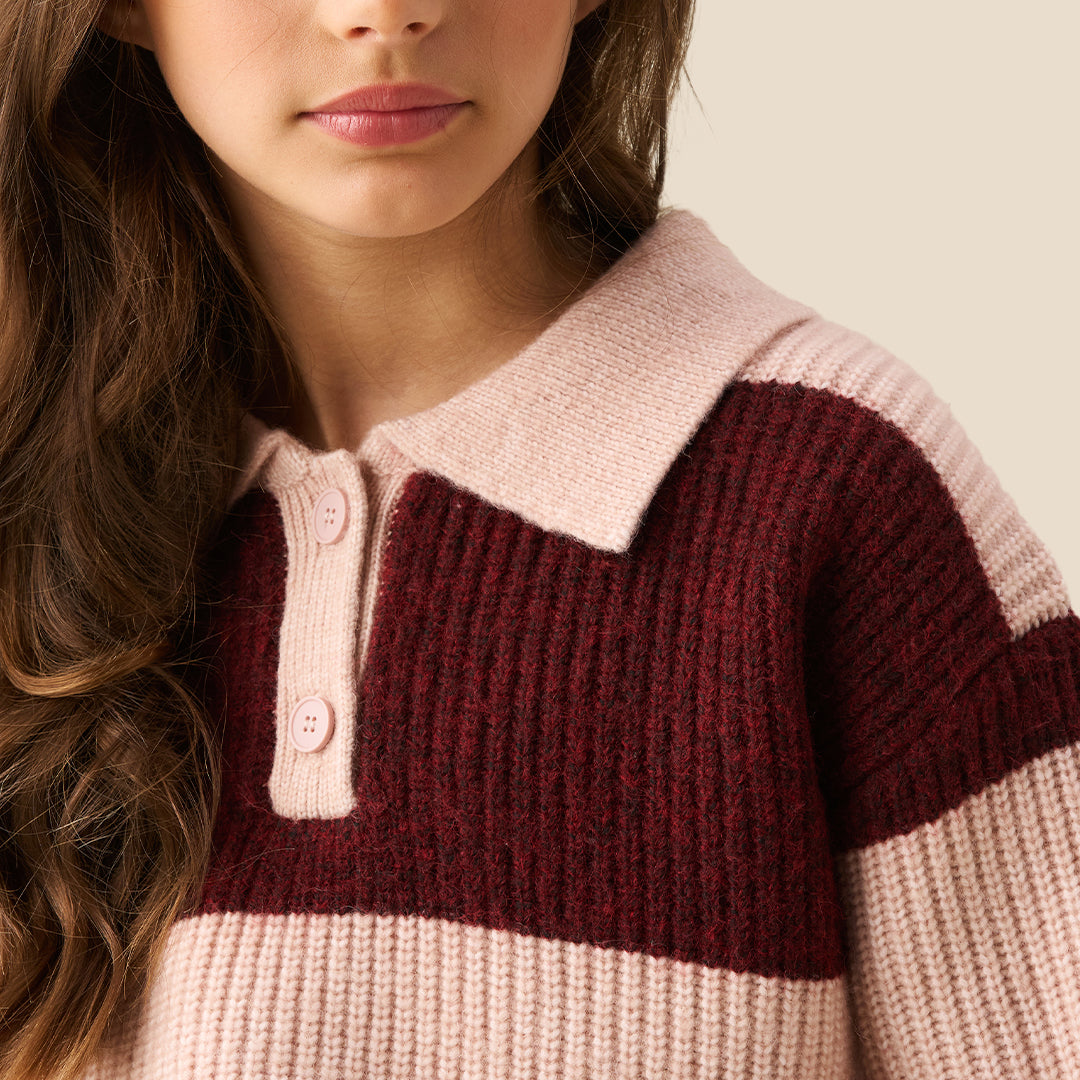 A young girl modeling a ribbed knit polo sweater with horizontal stripes in burgundy and light pink, paired with wide-leg blue jeans against a solid beige background.