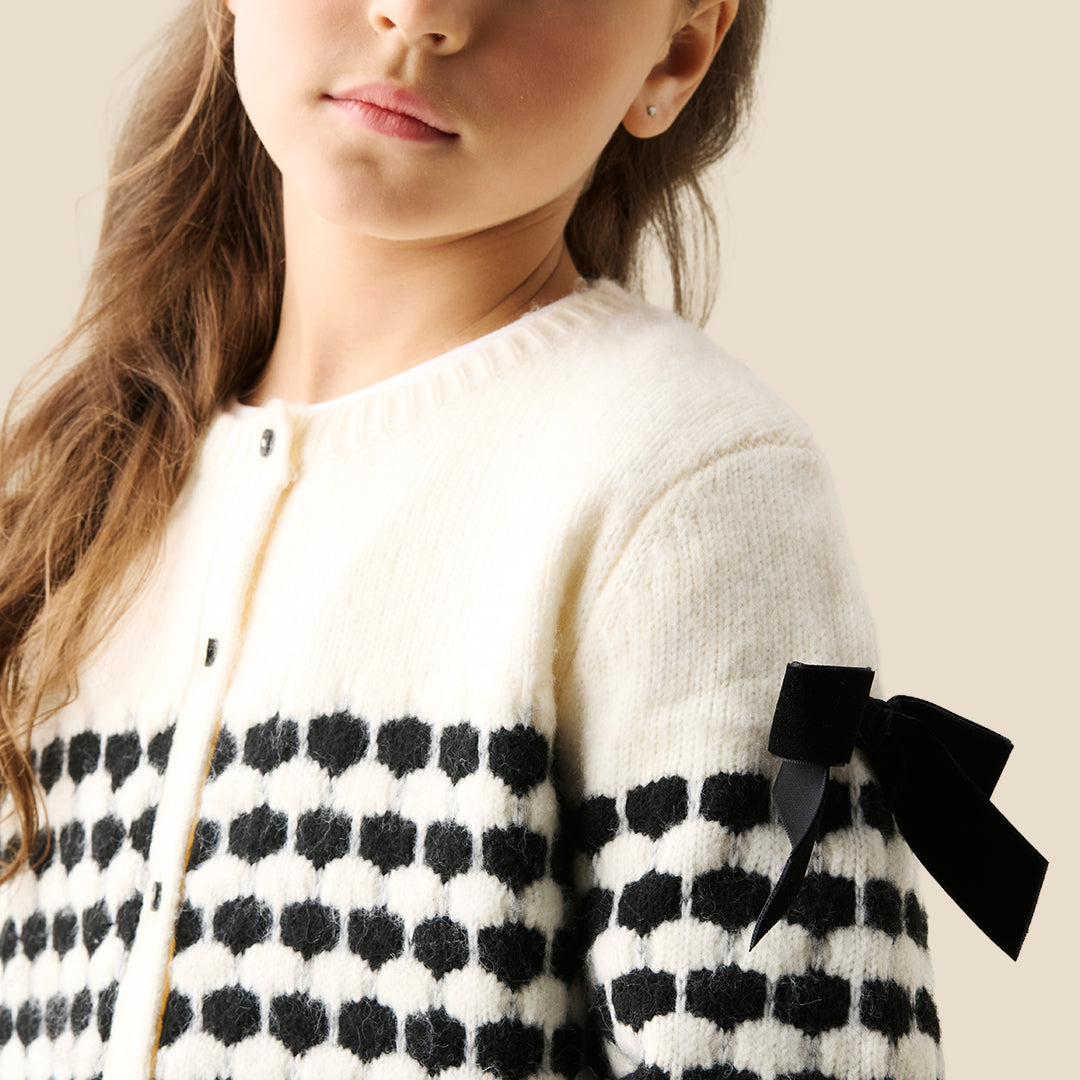 A young girl with her hair in a bun wears a cream knit cardigan with black polka-dot patterned sleeves and black ribbon bows on the shoulders, paired with a dark blue ruffled skirt, against a solid beige background.