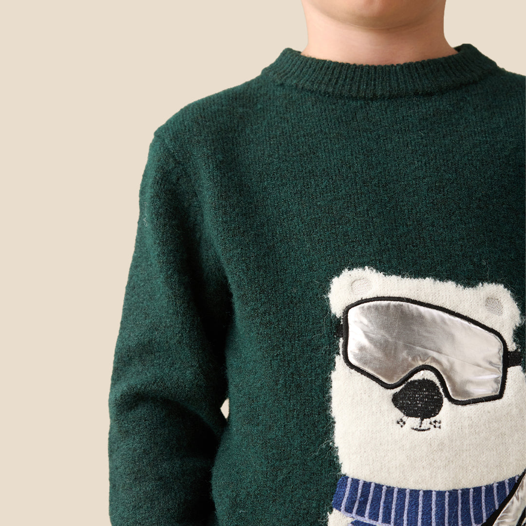 A young boy with blond hair wearing a dark green knit sweater featuring a large white polar bear graphic with ski goggles and a blue striped scarf, set against a solid beige background.