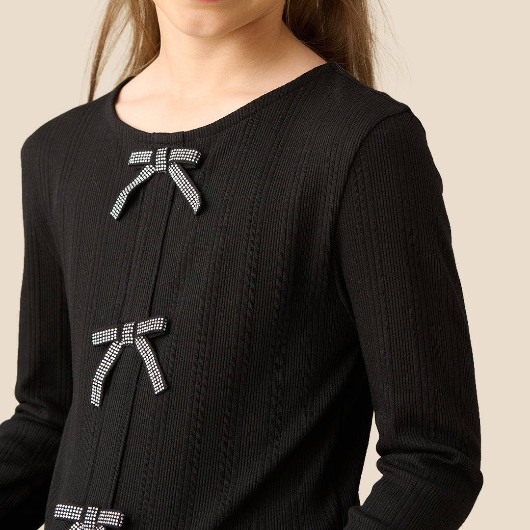 A young girl with light brown hair in a ponytail wearing a black long-sleeve ribbed top with three gray bow details on the front, paired with a black and white patterned skirt, against a solid beige background.