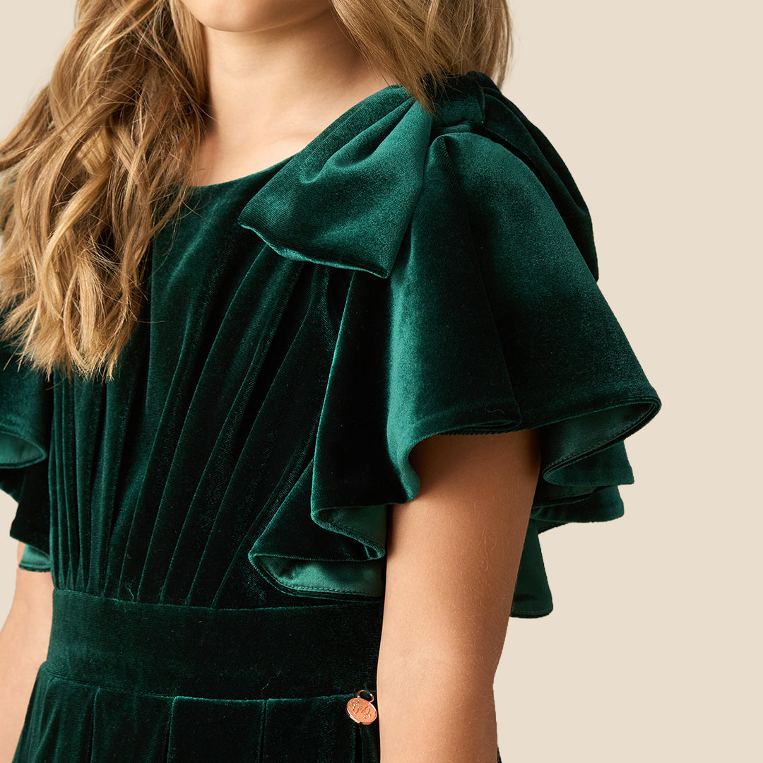 A young girl wearing a dark green velvet dress with ruffled short sleeves, a matching waist belt, and a pleated skirt, standing against a solid beige background.