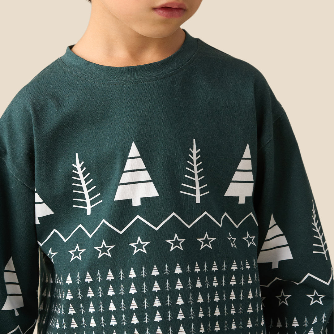 A young boy stands barefoot against a solid beige background, wearing a dark green long-sleeve pajama set with a white Nordic tree pattern.
