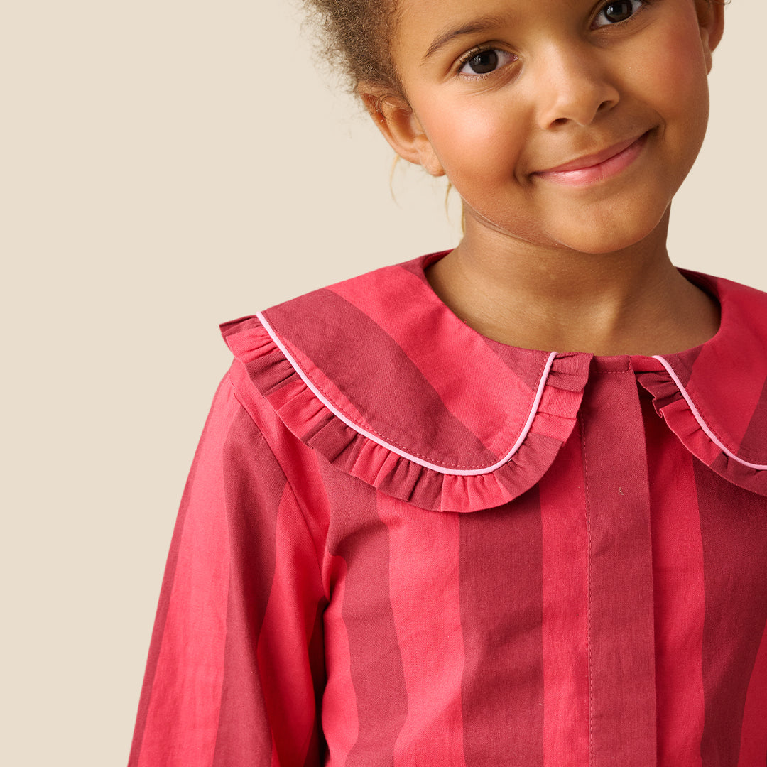 A young girl modeling a two-piece pajama set with vertical pink and red stripes, featuring a ruffled collar and white piping details, set against a solid beige background.