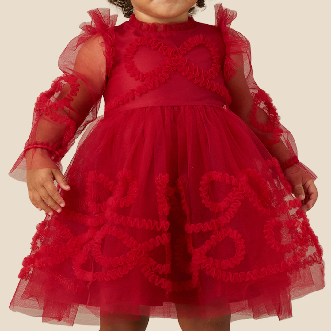 A toddler with curly brown hair wearing a red tulle dress with sheer long sleeves and red floral applique details, standing against a solid beige background.